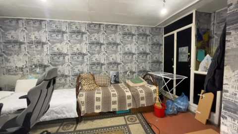 Media: Video of a small, cluttered bedroom with a single bed against the left wall, covered in a plaid blanket. The walls are adorned with black-and-white cityscape wallpaper. A white metal folding chair is beside the bed, and a red plastic bucket is on the floor.