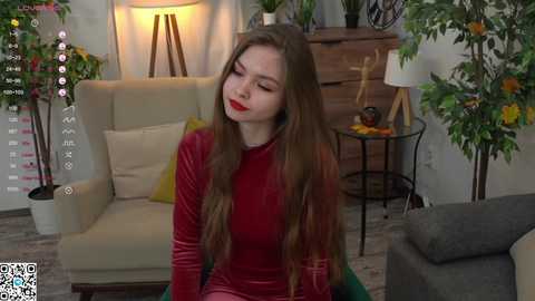 Media: Video of a fair-skinned woman with long brown hair, red lipstick, and a velvet red dress, sitting in a modern living room with green plants, wooden furniture, and a cozy lamp.