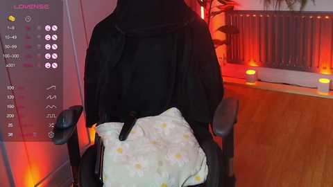 Media: Video of a person sitting in a black chair, wearing a black cloak, with a white blanket adorned with yellow polka dots, in a dimly lit room with warm red lighting.