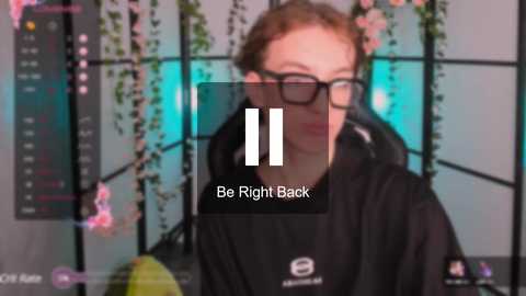 Media: Video of a person with glasses, light skin, and short red hair, wearing a black hoodie, in a dimly lit room with hanging plants and a digital clock. Text overlay reads, \"Be Right Back.\