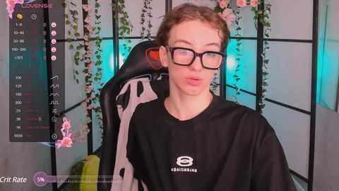 Media: A video of a young man with fair skin, curly brown hair, and black-rimmed glasses, wearing a black \"EBAKALIN\" shirt, seated in a black gaming chair with pink flowers and greenery in the background.