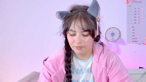 Media: Video of a young woman with long dark hair, wearing a blue cat ear headband, pink jacket, and \"MOM\" t-shirt, sitting in a soft pink-lit room with a calendar and dreamcatcher on the wall.