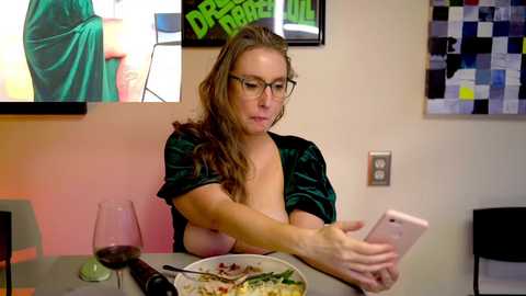 Media: Video of a white woman with glasses, long brown hair, wearing a green velvet top, revealing large breasts, sitting at a table, eating pasta, holding a phone, in a modern room with a TV, art, and wine glass.