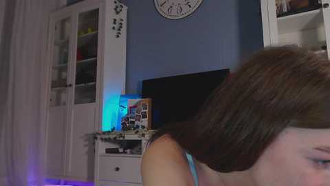 Media: Video of a young woman with shoulder-length brown hair, wearing a sleeveless top, in a dimly lit room with a TV, white shelves, and a wall clock.