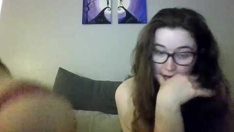 Media: A video shows a young woman with long brown hair, glasses, and a thoughtful expression, resting her chin on her hand. She's in a room with a gray couch, purple abstract wall art, and dim lighting.