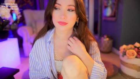 Media: Video of a fair-skinned woman with long brown hair, wearing a blue-striped shirt and red lipstick, posing seductively in a cozy, dimly-lit living room with soft lighting.