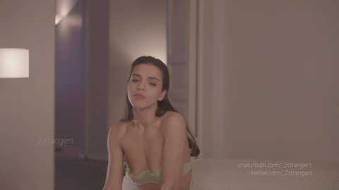 Media: A video of a young woman with long brown hair, wearing a strapless floral dress, leaning against a doorway in a dimly lit, minimalist room.