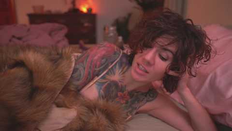 Media: Video of a woman with curly hair, tattoos, and a floral blouse, lying on a bed with a fur blanket, in a dimly lit bedroom with a lit candle.