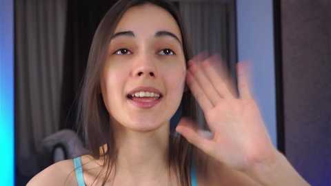 Media: A video of a young woman with light brown skin, straight dark brown hair, and a friendly smile, wearing a light blue top. She waves her right hand in front of a blurred, modern indoor background with blue and beige tones.