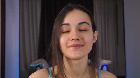 Video of a smiling, young woman with straight, shoulder-length brown hair, wearing a teal tank top, eyes closed, in a softly lit, dimly lit room with blurred curtains in the background.