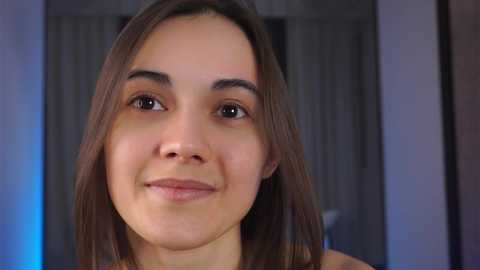 Media: A video of a young woman with medium-length, straight brown hair, light skin, and a soft smile. She has well-defined eyebrows and wears minimal makeup. The background features blurred curtains and a blue light source.