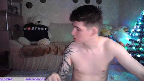 Media: Video of a shirtless young man with a black tattoo sleeve, sitting indoors next to a decorated Christmas tree, surrounded by plush toys and a beige couch.