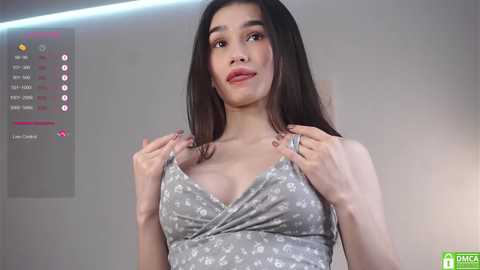 A video of a young woman with long dark hair, wearing a gray floral-patterned dress, standing in a minimalist room.