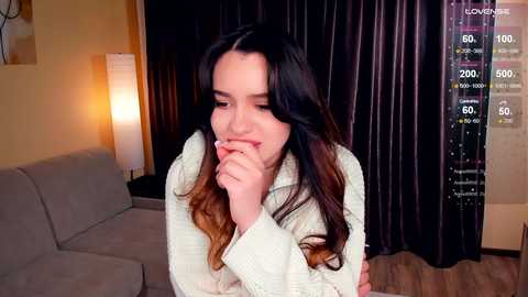 Media: A video of a young woman with long, dark hair, wearing a beige sweater, sitting on a sofa, looking thoughtful, in a dimly lit living room with dark curtains.