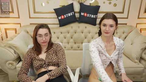 Video of two young women sitting on a beige tufted sofa in a modern living room. One wears a leopard print top, the other a floral cardigan, both with glasses. Black pillows with white text read \"Mastbet\" on the back wall.