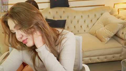 Video of a young woman with long, wavy brown hair, wearing a light sweater, leaning on a beige, tufted sofa with beige and black pillows, in a cozy living room.