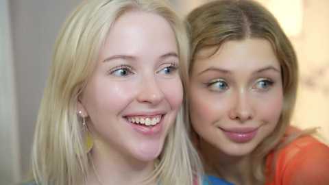 Media: Video of two young women with fair skin and blonde hair, smiling and laughing. One wears a yellow necklace, the other has a red top.