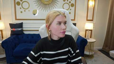 Video of a blonde woman in a black and white striped sweater, seated on a white chair in a modern living room with blue couches, gold-framed mirrors, and decorative lamps.