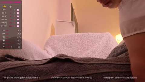 Media: Video of a person's torso in a cozy bedroom, wearing a white shirt, standing next to a gray and white textured bedspread. Background includes a white nightstand and warm, soft lighting.