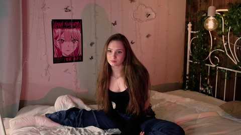 Media: Video of a young woman with long brown hair, sitting on a bed in a dimly lit room with pink walls, wearing a black top and jeans, surrounded by floral decorations and a metal bed frame.