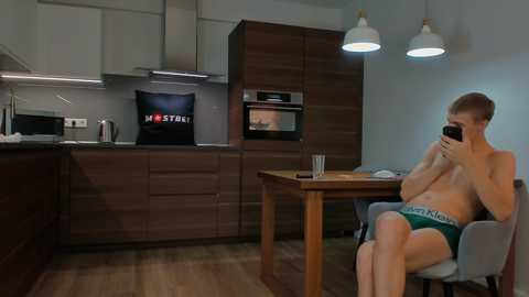 Media: Video of a young, shirtless man with fair skin and short blonde hair, wearing green Calvin Klein briefs, sitting at a wooden table in a modern kitchen with wooden cabinets and stainless steel appliances.