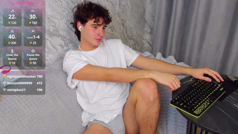 Media: Video of a young, curly-haired man in a white T-shirt, shorts, and headphones, sitting on a grey couch with a gaming keyboard.