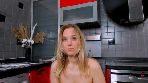 Media: Video of a topless Caucasian woman with long blonde hair sitting in a modern kitchen with gray tiled walls, black countertops, and red and silver cabinets.