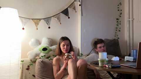 A video of a nude woman with long hair sitting on a beige couch, holding a mug, in a cozy, dimly-lit room with a stuffed toy, a lamp, and a computer.