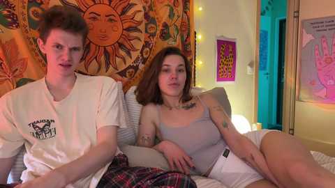 Media: Video of two women, one with short hair and the other with long hair, sitting on a bed in a cozy room with a sunburst tapestry and colorful artwork.