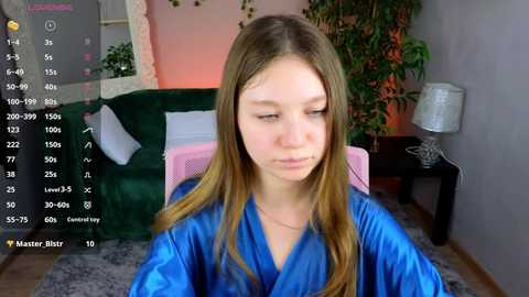 Media: A video of a young woman with long, straight, light brown hair in a blue satin robe, seated in a cozy room with green velvet furniture, a potted plant, and a lamp.