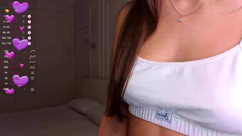 Media: Video of a woman with light skin, wearing a white crop top, long brown hair, and a necklace, in a dimly lit bedroom with hearts overlay.