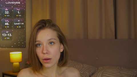 Media: Video of a young woman with shoulder-length brown hair, blue eyes, and fair skin, sitting on a bed with a patterned blanket. Background features a dimly lit room with a lit lamp, a calendar, and a window with beige curtains.