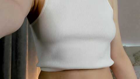 Media: Video of a fair-skinned woman wearing a white ribbed tank top, highlighting her slender waist and small breasts. Background shows a blurred, neutral-toned room.