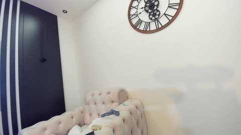 Media: Video of a minimalist bedroom with a beige tufted sofa, a large round clock on the wall, and a dark blue curtain partially visible in the background.