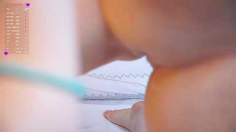 Video of a close-up view of a person's hand holding a pen, writing on a white pad with zigzag lines. The background shows a blurred, light-colored room with a soft, warm lighting.