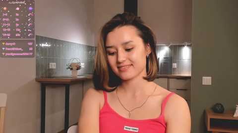 Media: Video of a young woman with light skin, straight brown hair, wearing a pink tank top, in a modern kitchen with tiled backsplash and minimalistic decor.