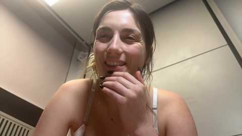 Media: Video of a young woman with wet hair, wearing a white tank top, holding a phone to her ear, smiling, in a bathroom with beige walls and a radiator.