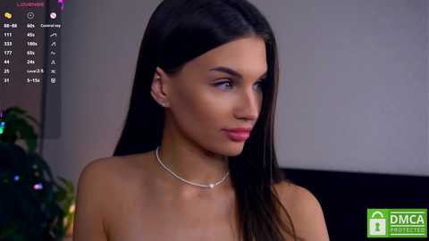 Media: Video of a topless woman with long, straight black hair, wearing a silver necklace, in a dimly lit room with a TV screen and green logo.