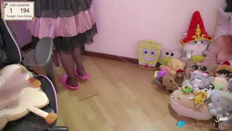 Media: Video of a young woman in a black dress with pink tulle and pink shoes standing near a plush toy collection on a wooden floor.