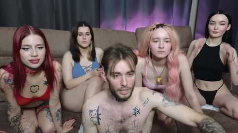 Media: Video of five young people, including a tattooed man, sitting on a couch in a dimly-lit room. They wear revealing clothes, with the man shirtless.