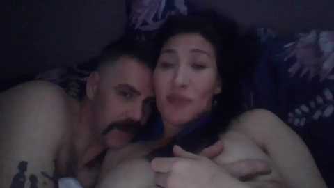 Media: Video of a naked man with a beard and a naked woman with dark hair, both smiling, lying on a bed with floral-patterned sheets in dim lighting.