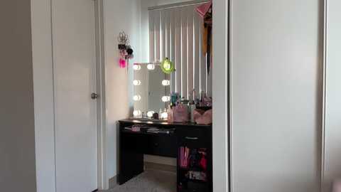 Media: Video of a well-lit, organized vanity with a large, rectangular mirror adorned with bright bulbs, pink and black decor, and a black desk cluttered with makeup items.