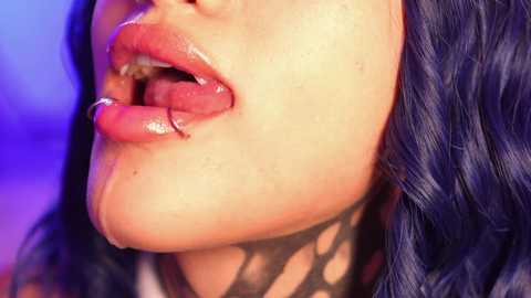 Media: Video of a woman with light skin and blue hair, her mouth open with a shiny, white substance on her lips and tongue, wearing a black fishnet top. Background is a blurred mix of blue and purple hues.