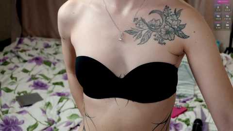 Media: Video of a light-skinned person with a small chest, wearing a black strapless bra, displaying a detailed floral tattoo on the right shoulder. The background features a bed with a floral-patterned sheet and scattered items.