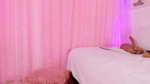 Media: Video of a minimalist bedroom with pink curtains and white bed, featuring a nude person lying on the bed, with a pink stool beside the bed.