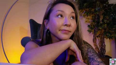 Media: Video of an Asian woman with long brown hair, wearing a sleeveless top, resting her chin on her hand. She has a serene expression and a tattoo on her arm. Background includes a yellow wall, green plant, and a modern chair.