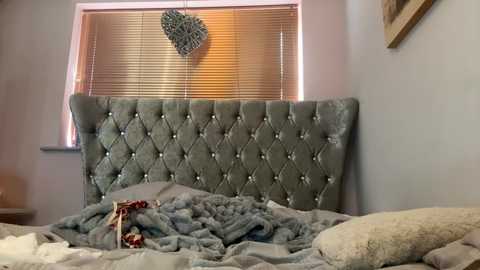 Media: Video of a cozy bedroom with a plush, tufted, grey velvet headboard, soft grey blankets, and a heart-shaped wall decoration above a window with closed blinds.