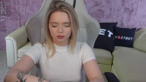 Media: Video of a young Caucasian woman with blonde hair, medium build, wearing a tight white top, sitting in a white gaming chair with a purple wall and \"MST BET\" pillow behind her.