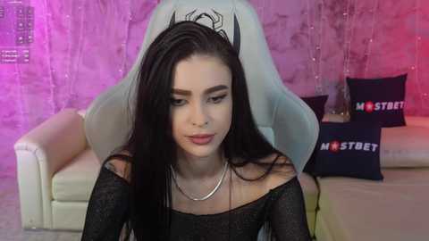 Media: Video of a young woman with long black hair and pale skin, wearing a black off-shoulder dress and silver necklace, seated in a white gaming chair in a room with pink and purple lighting, surrounded by \"MOSTREET\" pillows.