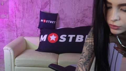 Media: Video of a tattooed woman with long black hair and pale skin, wearing a sleeveless top, sitting on a cream sofa with a dark navy pillow featuring \"MOSTB\" text and a red star. Background is a textured, purple wall.